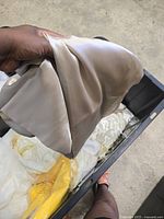 Hand holding a light cream satin tablecloth with shiny, smooth texture above a black plastic bin containing more tablecloths.