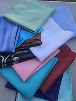 Photo shows a stack of assorted fabric table napkins in various colors: light green, light blue, turquoise, pink, white, brown, maroon, black with white design, and dark blue.