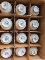 View of 36 white ceramic teacups, bottom markings visible, organized in cardboard box compartments.