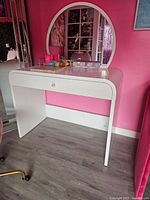 White vanity desk against pink wall, showing large round mirror and single central drawer with a clear knob, placed on laminate flooring.