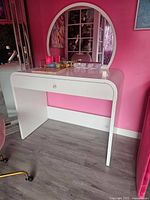 White wooden vanity set against a bright pink wall. Oval mirror attached to table back. Table has single drawer with knob. Matching pink cushioned stool with gold legs on wheels is partially visible.
