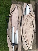 Top-down view of two beige carrying bags open showing collapsed white metal frames and partially folded beige canopy fabric.