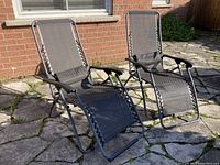 Two black anti-gravity lounge chairs positioned outdoors on a stone patio showing general wear and damage to plastic armrests.