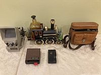 Photo showing a vintage Dual 8 film reviewer in gray, a decorative train bottle holder with shot glasses, a vintage video camera in a brown leather case, a small black case, and a small black electronic device with a red button.