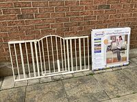 Photo showing white metal baby gate with vertical bars, 6 feet wide, leaning against a brick wall with original product box on right.