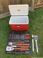 Open red Coleman cooler with white interior next to a black BBQ utensil case with wooden-handled tools and two additional stainless steel utensils with metal handles