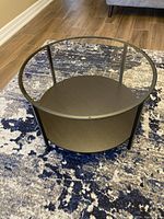 Photo showing the round coffee table with a clear glass top and dark solid base, placed on a patterned rug.