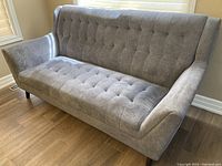 Full frontal view of blue fabric couch showing tufted back and seat cushions with angled arms and wooden legs on hardwood floor.