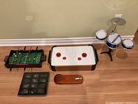 Photo showing the full lot with all items: tabletop foosball table, air hockey table, mini drum set, boxed parlour puzzles, and mini bowling alley game on a wood floor with a beige wall background.