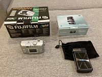 Four cameras shown with some original boxes: Fujifilm 1400Zoom, Canon ELPH135, Insignia camera visible with pouch.