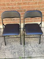 Front view of two black folding chairs placed side by side on pavement against a brick wall.
