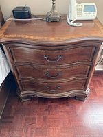 Front view of the 3-drawer wooden nightstand showing curved drawer fronts and metal handles, with some minor surface scuffs visible on the top.