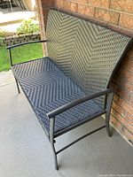 Front angle view of black woven wicker outdoor bench with metal frame against brick wall on concrete.