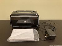 Photo of HP photo printer along with power adapter and plastic bag of photo sheets on dark surface