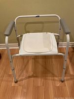 Portable toilet seat closed with padded armrests on each side. White metal frame with adjustable legs.