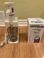 Cuisinart hand blender standing in a clear plastic measuring/chopping cup next to the Proctor Silex electric can opener in its box on a hardwood floor.