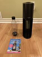 Black Sodastream drink maker with clear plastic carbonating bottle and instruction booklet on wooden floor.
