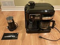 DeLonghi espresso machine and Aeroccino 3 milk frother placed on floor with power cords visible. Both items shown from front.