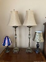 Three lamps on a wooden surface including two matching tall brass lamps with pleated shades, a smaller Tiffany style stained glass lamp, and a smaller decorative lamp.