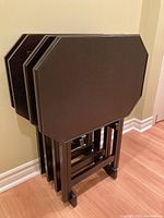 Front side view of four wooden TV trays stacked in the wooden stand, showing the trays' flat top surfaces and the stand's structure.