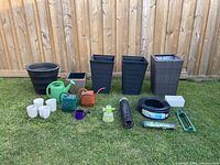 Photo showing 5 large planters in black and brown, 5 white small ceramic planters, 4 watering cans in various colors, green handheld garden spray, lawn edging roll, weed control fabric roll, and drainage pipe spread on grass.