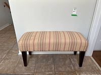 Full view of the upholstered bench with beige striped fabric and dark wood legs on a tile floor.