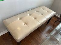 Full view of the rectangular ivory bonded leather bench showing the button tufted upholstery and dark wooden legs angled outward.
