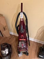 Front view of the burgundy Bissell ProHeat Pet carpet cleaner with hose and upholstery nozzle attached, along with a bottle of cleaning solution placed on the floor.