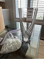 Clear cut crystal vase, bag of transparent glass pebbles on glass table beneath window light.