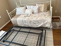 Metal day bed with bedding and mattress, decorative pillows arranged on the bed, and folded trundle bed frame in front of the bed.