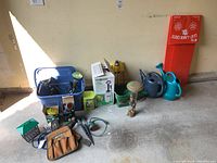 Wide view of assorted gardening supplies including watering jugs, a box sprayer, garden stake lights, tool pouch, and bird feeder on a concrete floor.