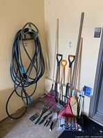 Overall view of gardening tools set including hose rack, two shovels, rake, hoe, hand tools, dustpan, and garden soil bags.