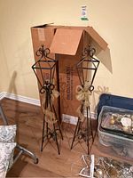 Pair of tall wrought iron candle holder lanterns standing on floor by a cardboard box and plastic bin.