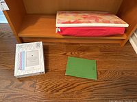 Kobo E-Reader in green cover, Kobo packaging box, and Indigo lap desk with colorful top and red base, on wooden floor and shelf.