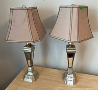 Two reflective silver lamps with beige fabric shades, tri-light sockets on display on table.