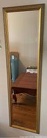 Full view of the tall, narrow rectangular wall mirror leaning against a wall, showing gold decorative frame and clear reflective surface.