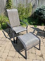 Photo shows metal and plastic mesh reclining patio chair and matching ottoman outdoors on stone patio with greenery background.