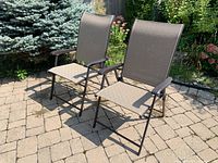 Pair of folding patio chairs shown on a paved surface with a garden background. Chairs are metal framed with brown plastic mesh seats and backs, and black plastic armrests.