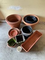 Overview image showing all planters arranged on the floor, including large clay pots, black plastic pots nested inside, rectangular planter, and small decorative pots in various colors and shapes.
