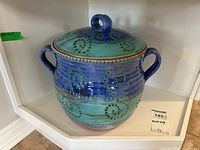 Front angle view of the lidded pottery crock showing the blue and green glaze with circular patterns and dual handles.