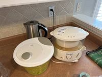 Three kitchen appliances on countertop: stainless steel electric kettle, white electric steamer, and salad spinner with green basket.