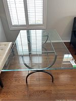 Top view of rectangular clear glass table showing black curved metal base underneath and hardwood flooring.