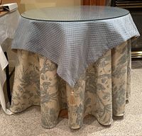 Full view of round wooden framed table with a clear round glass top and layered fabric covers including a blue quilted cloth and a beige floral patterned skirt underneath with tassel detail.