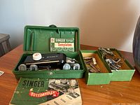 Vintage Singer buttonholer in green case with templates and instruction booklet, plus parts box on table.