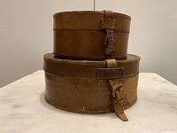 Front view of the two antique British leather collar boxes stacked, showing the taller box on top with a leather strap and buckle closure, and the wider box underneath with worn leather and a buckle.