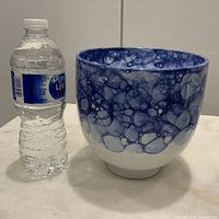 Front view of the blue and white footed planter next to a small water bottle for size comparison, showing bubbly glaze pattern and footed base on a white background.