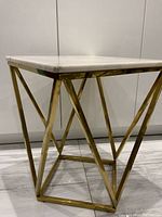 Close angled view showing square white marble top with slight wear and a small chip on one corner, sitting on a polished brass geometric base.