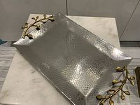 Photo of hammered stainless steel rectangular tray with gold gilt vine leaf handles on a white marble surface.