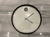 Front view of the round wall clock showing white face, black hands and signature black dot at 12 o'clock.