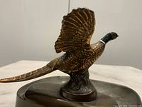 Side angle of bronze bird figurine mounted on the rim of the coin tray, showing details of the bird's feathers and overall condition.